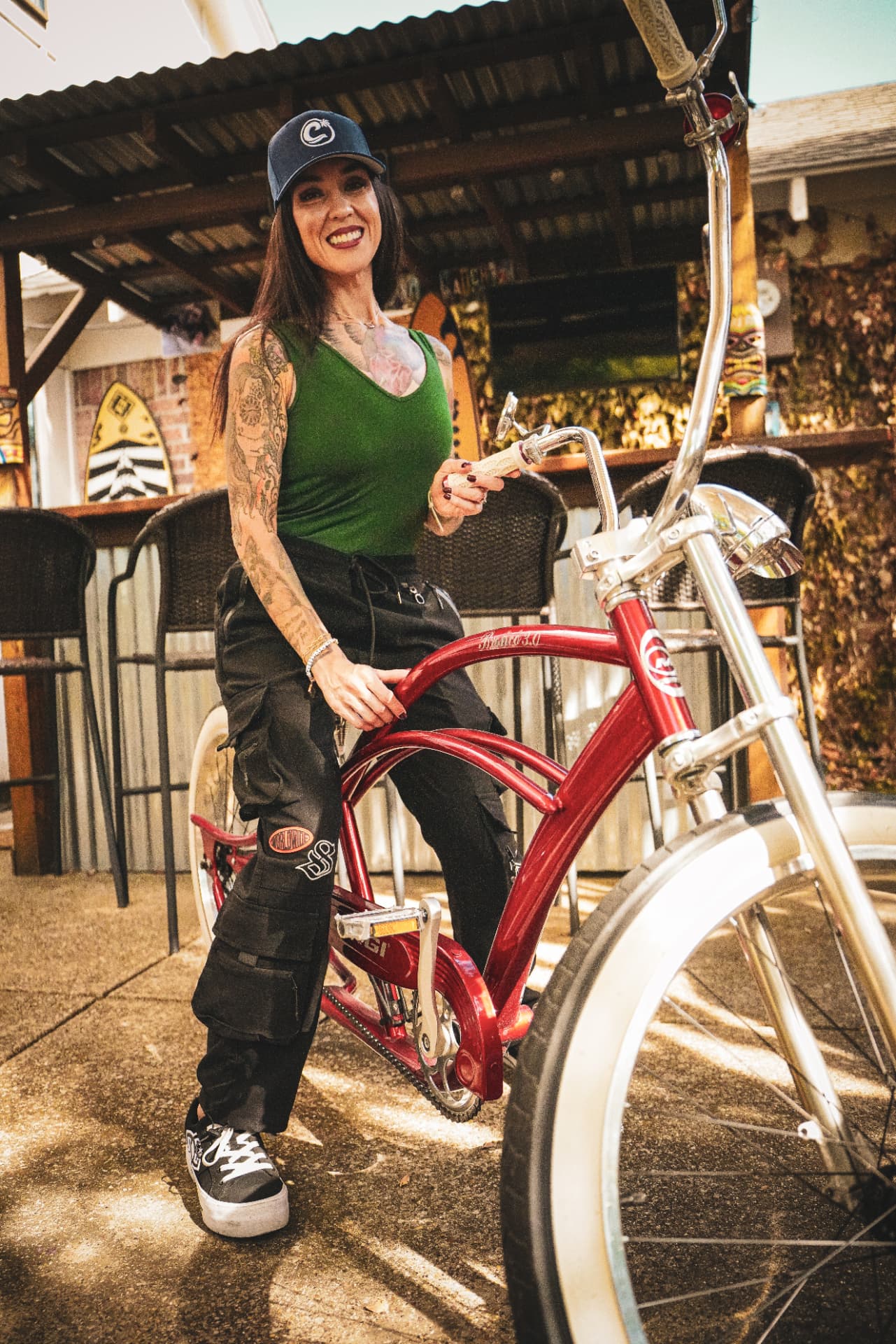 Dani smiling at the camera in a navy Castaway Shades cap beside a red cruiser bike.